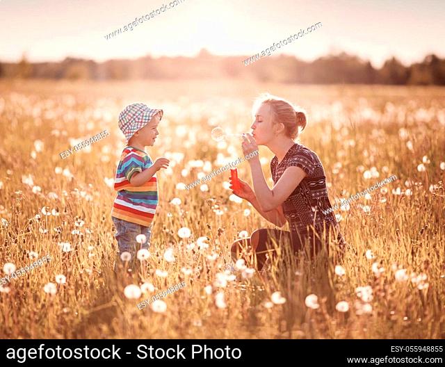 Young woman with a boy blowing bubbles. Mother and son having fun on dandelion field