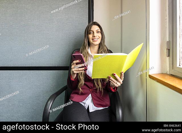 Young blonde business woman sitting in her office using the phone while checking notes looking at the camera