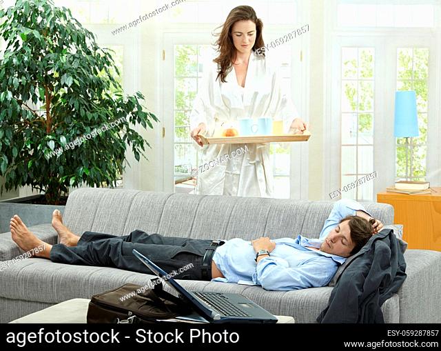 Tired businessman resting on couch at home in the morning, his wife brining breakfast on tray