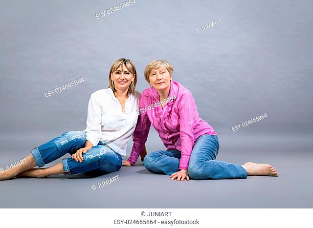 Attractive stylish blond senior lady with her beautiful middle-aged daughter posing together with her hands on her shoulders smiling at the camera on a grey...