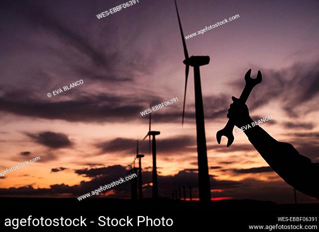 Silhouette of engineer holding wrench by wind turbines at wind farm on sunset