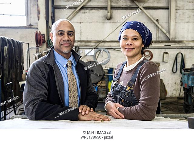 Black man owner of a sheet metal factory and a black woman factory worker going over plans at a work station on the floor of the factory