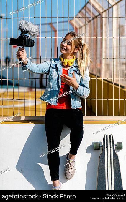 Smiling blond woman recording video through camera while sitting on surrounding wall