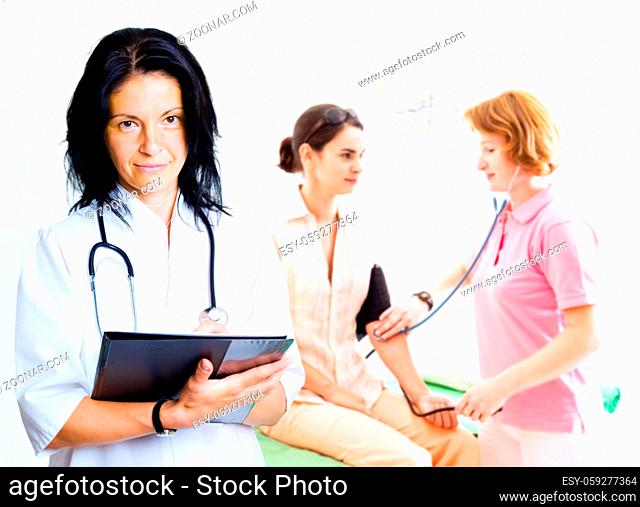 Young female doctor is looking up at the camera while nurse is measuring patient's blood pressure in the background