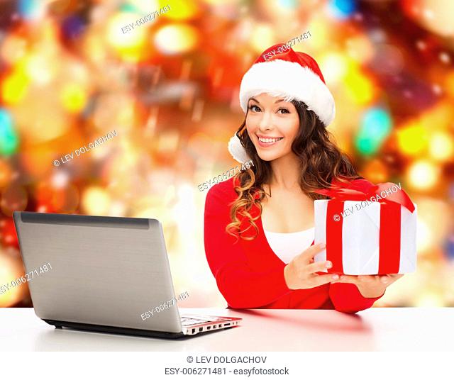 christmas, holidays, technology and people concept - smiling woman in santa helper hat with gift box and laptop computer over red snowing background