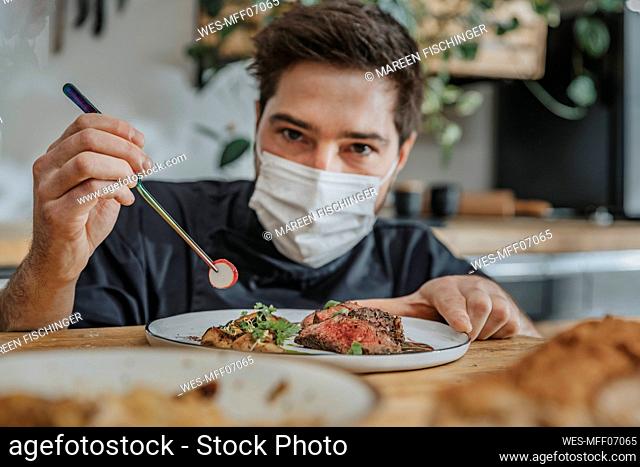 Male chef wearing protective face mask garnishing vegetable on tomahawk steak while working in kitchen