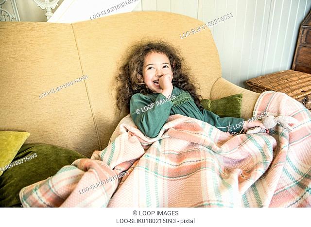 A six year old girl holding a toy rabbit sitting on a sofa wrapped in a blanket