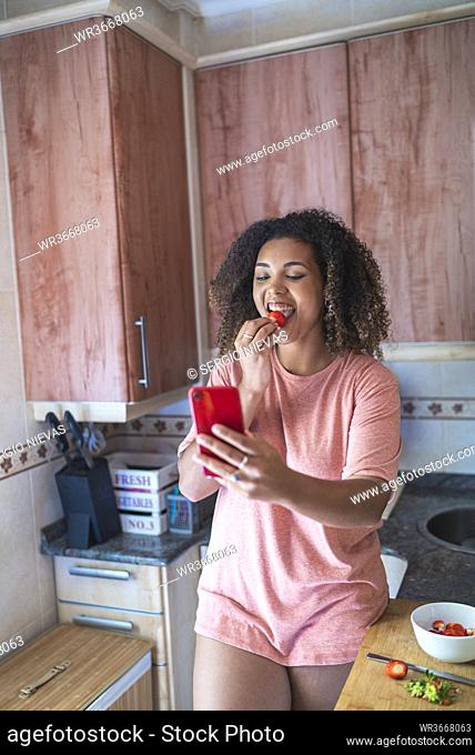 Cheerful young woman eating strawberry while video calling over smart phone in kitchen