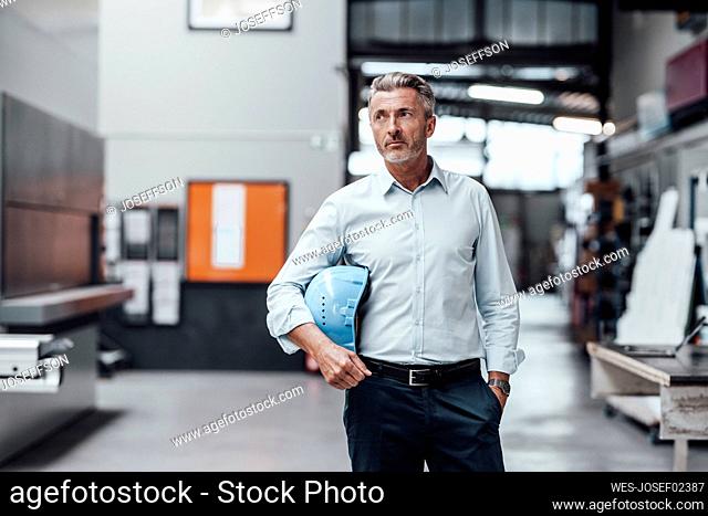 Thoughtful male engineer holding hardhat while standing in factory