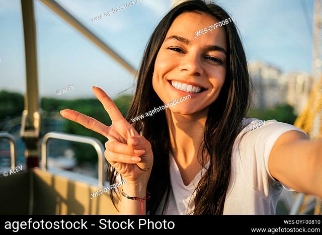Happy woman showing peace gesture and taking selfie in Ferris wheel
