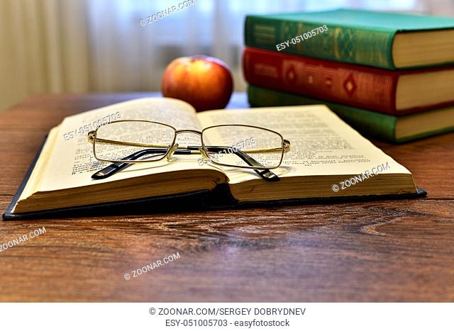 Open book with glasses, apple and stack of books on the table