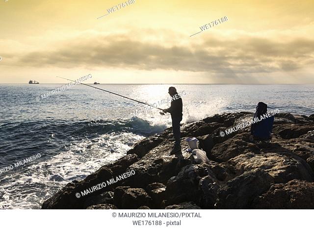 Savona, Italy. A fisherman standing with the fishing rod in his hand, he is on the rocks at sunset, near him buckets and equipment in bags