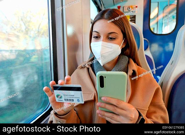 Portrait of young woman with protective face mask traveling with train paying with her credit card on smart phone for shopping online