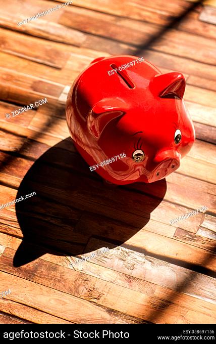 Ein rotes Sparschwein steht auf einem Holzboden und wirft einen starken Schatten A red piggy bank stands on a wooden floor and casts a strong shadow