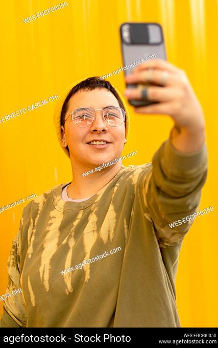 Happy non-binary person taking selfie through mobile phone in front of yellow corrugated wall