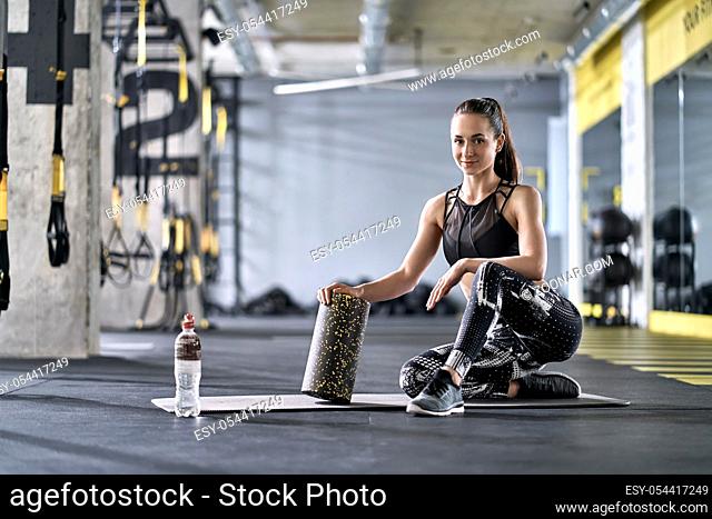 Smiling girl is posing with a black-yellow foam roller on the gray mat in the gym. She wears a black top with dark pants with prints and gray sneakers
