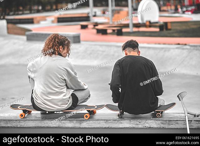 Good time. Back view of girl with long wavy hair and guy with crutches sitting on skateboards talking in park