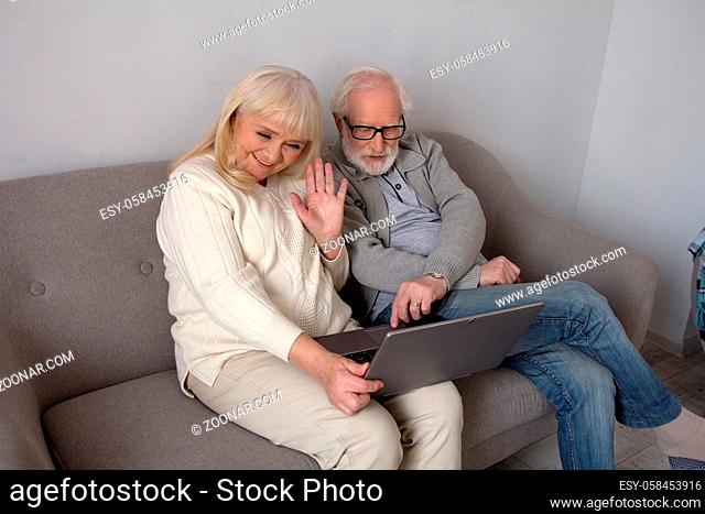 Old grandparents talking to their children via computer on sofa in living room. Beautiful aged lady waving at laptop next to her husband on couch