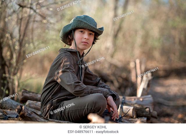 A boy in a coveralls and a hat sitting on a pile of sawn logs in the forest