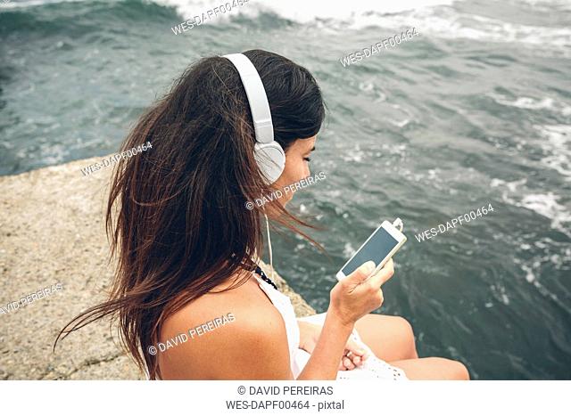 Woman listening music with headphones looking at smartphone in front of the sea