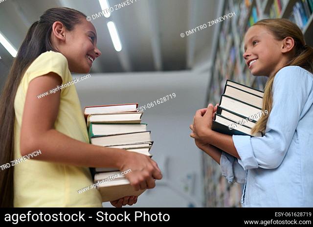 Two cheerful schoolgirls with stacks of books in their hands smiling at each other in the library