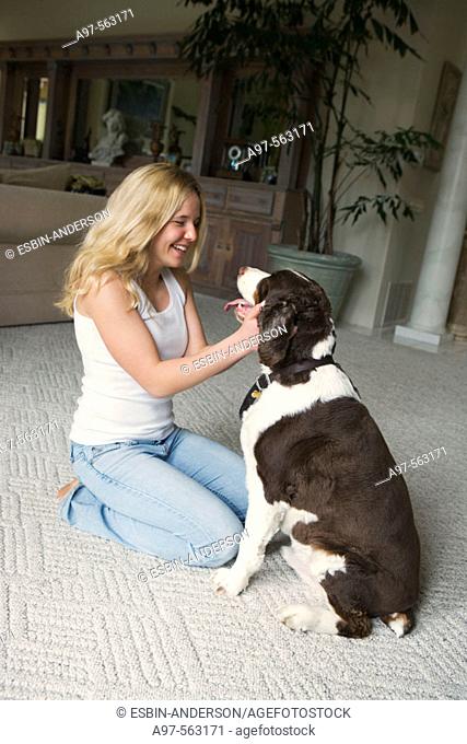 Smiling blonde teen girl in blue jeans and white tank top kneeling, caressing her pet dog's face
