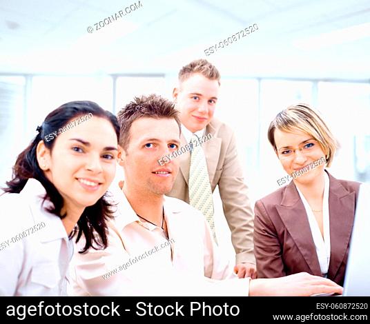 Four business colleagues working together on laptop computer in office, looking at camera, smiling. Focus placed on sitting man in middle