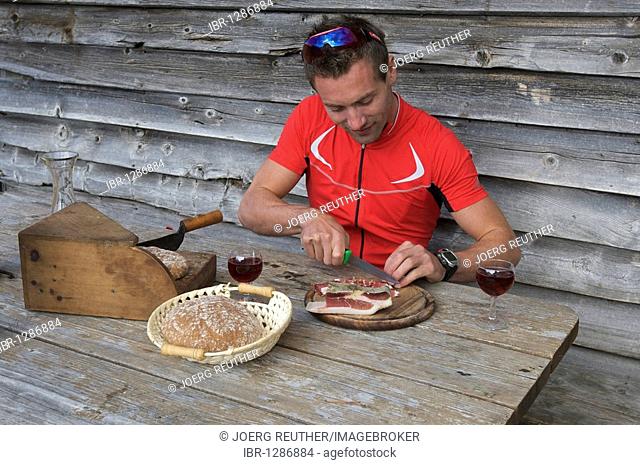 Bicycle racer cutting South Tyrolean bacon at the mountain lodge on the Passo di Pennes, South Tyrol, Italy, Europe