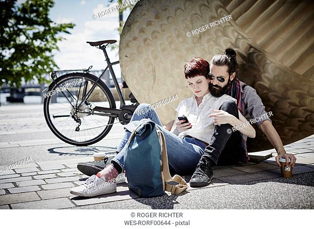 Germany, Hamburg, couple on bicycle trip having a rest looking at cell phone