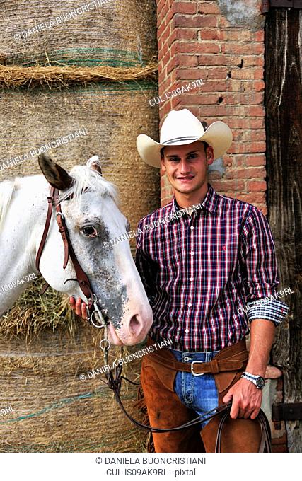 Portrait of young man in cowboy gear with horse outside stable
