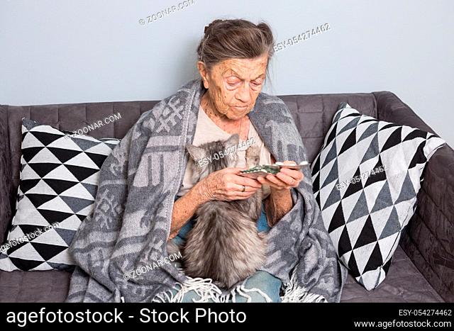 theme old age, loneliness health care. old grayhaired Caucasian woman with deep wrinkles sitting with pet animal cat. poor grandmother holding blister pill...