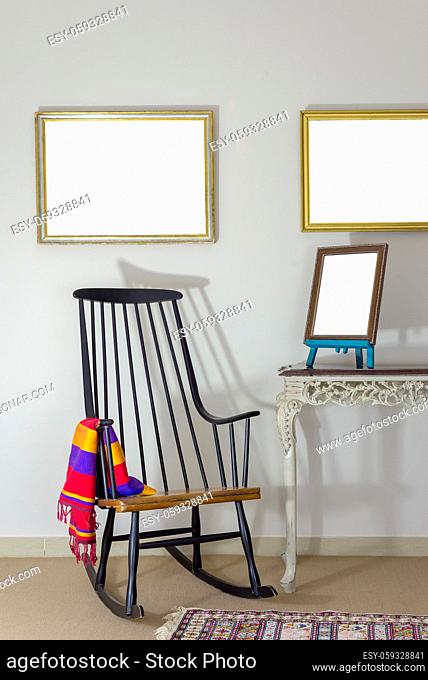 Interior shot of classic rocking chair and wooden ornate brown desktop photo frame on old style vintage table on background of off white wall with two hanged...