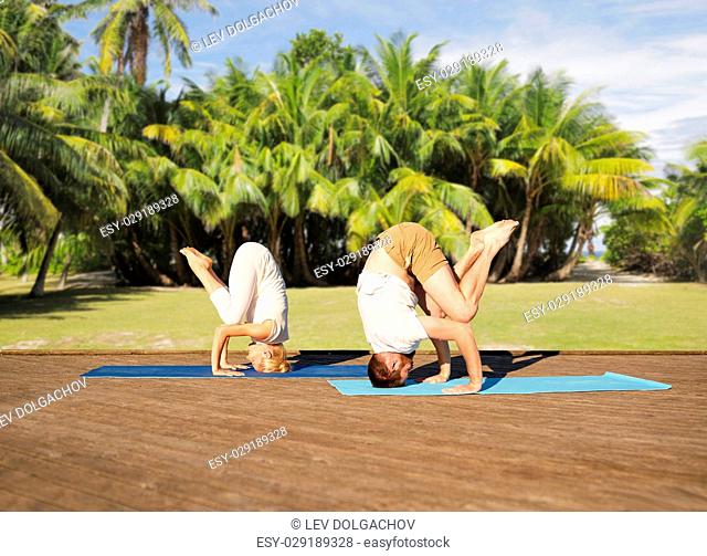 fitness, sport, yoga, people and healthy lifestyle concept - couple making headstand outdoors over natural exotic background with palm trees
