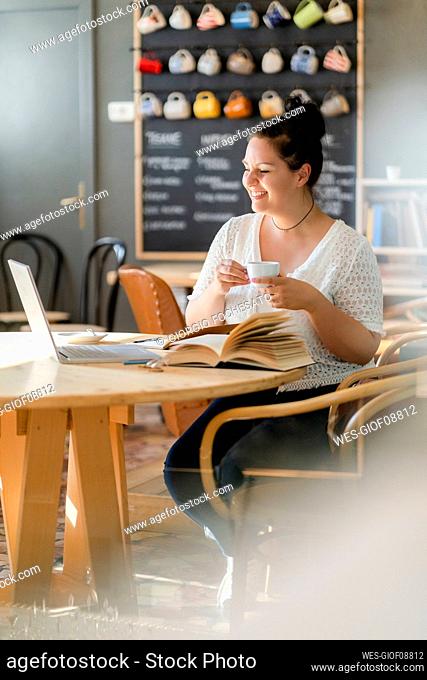 Smiling voluptuous woman holding coffee cup while studying over laptop in restaurant