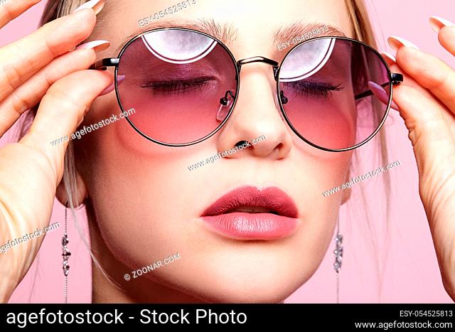 Close-up portrait of attractive young woman in tinted glasses and closed eyes. Female looking at camera on pink background