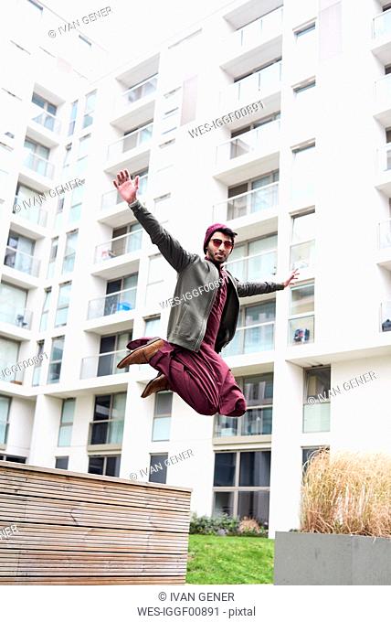 Full length of stylish man in purple hat and Indian costume jumping over modern building with widened arms
