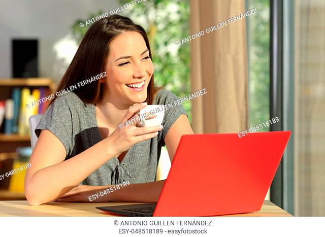 Happy woman with a laptop holding a cup of coffee relaxing and thinking looking through a window at home
