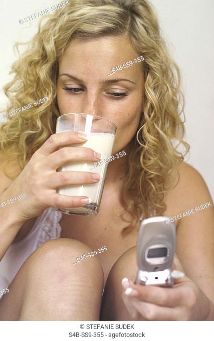 Junge blonde Frau mit Locken mit einem Handy in der Hand trinkt von einem Glas mit Milch - Morgens - Alltag , Young blonde Woman with curly Hair holds a Mobile...