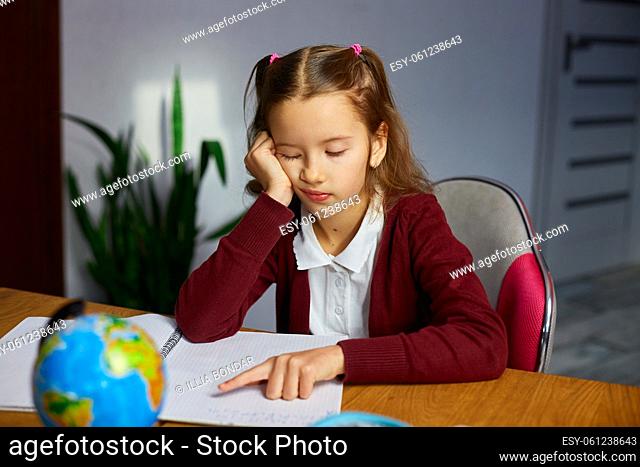 Bored schoolgirl sit at desk doing homework reading, homeschooling. Small junior girl tired of distant learning get remote elementary education and preparing...