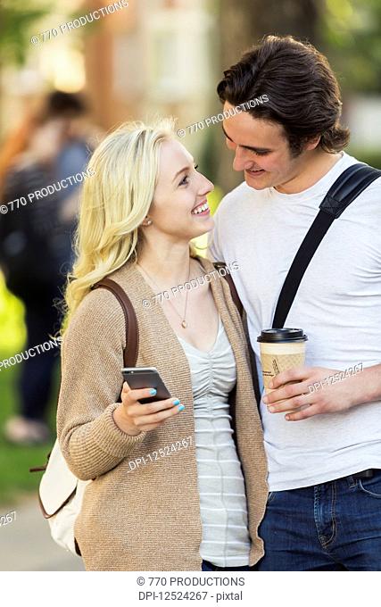 A young couple looking into each other's eyes and checking social media on a smart phone while walking through a university campus; Edmonton, Alberta, Canada