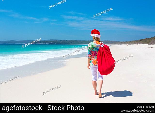 Christmas for a lot of Australians means hot summer sunshine and days spent at the beach. A woman wearing a santa hat strolls along a white sandy beach with a...