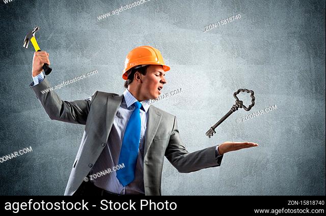 Furious businessman going to crash with hammer steel key. Young handsome contractor in business suit and safety helmet standing on grey wall background