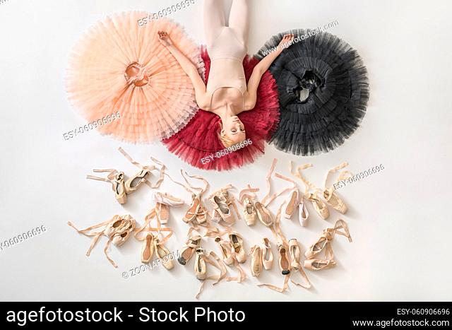 Gorgeous ballerina lies on the colorful tutus on the white floor in the studio. She wears light dance wear. Below her there are many pointe shoes
