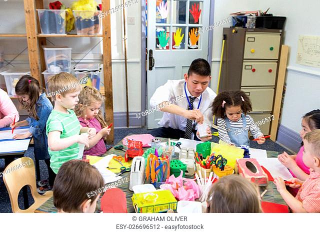 Male teacher with a class full of nursery students. They are all sat at tables using arts and crafts