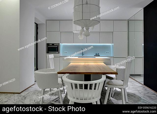 Modern kitchen with white walls and gray tiles with patterns on the floor. There is a wooden table with white chairs, kitchen island with a stove, white lockers