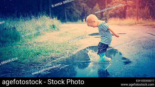 Child walking in wellies and jumping in puddle on rainy weather. Boy under rain in summer outdoors