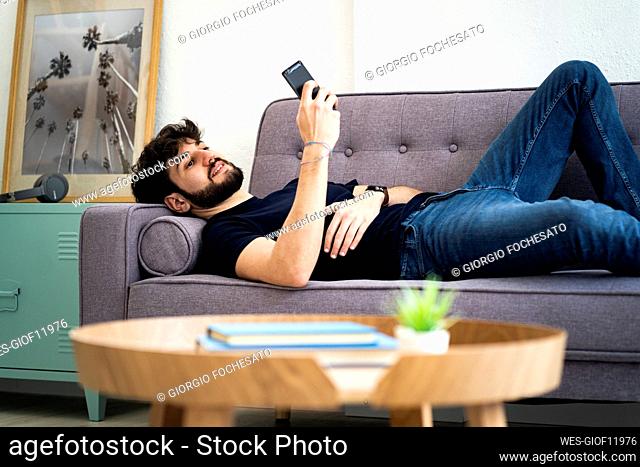 Smiling man using smart phone while relaxing on sofa in living room at home