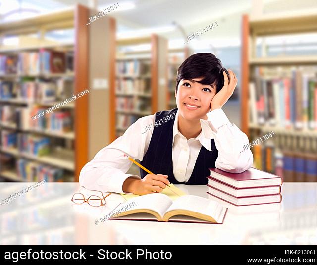 Young female mixed-race student with books and paper daydreaming in library looking to the left