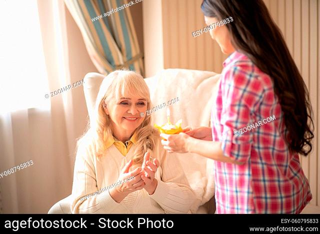 Delighted Elderly Mother Takes Her Birthday Muffin With Burning Candle From Her Daughter's Hands. Brunette Daughter Celebrates Her Old Mother
