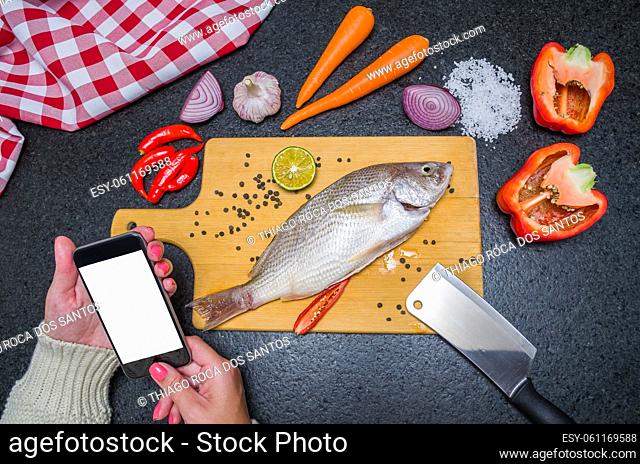 Young woman consulting recipe for fish on smartphone, fish and ingredients on the table. Preparing a gastronomic dish
